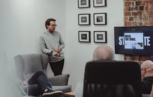 A man in a gray hoodie stands by a gray armchair, presenting to a group seated in a